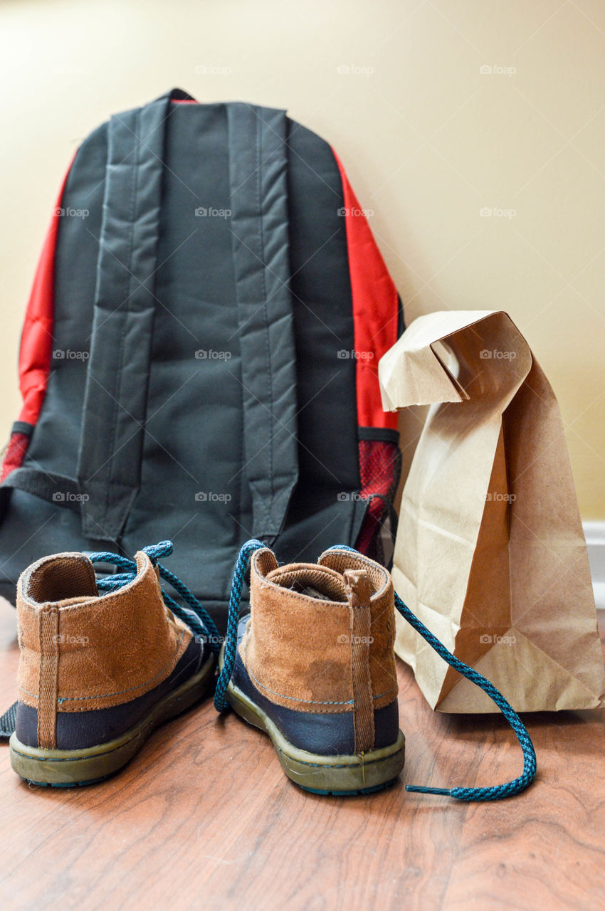 Child's backpack, brown bag lunch and shoes sitting on a wooden floor