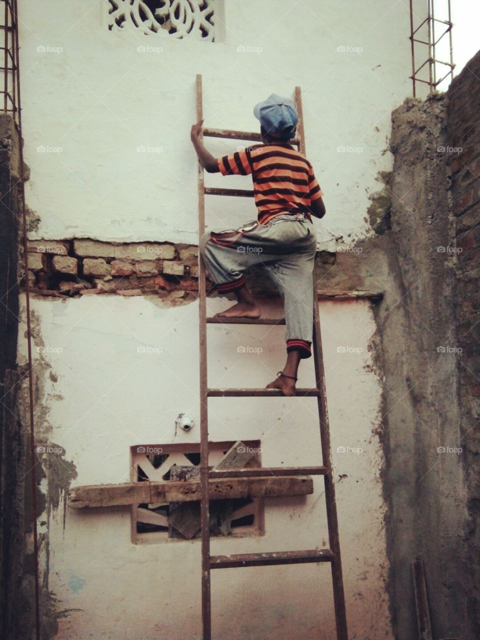 Photographs of a child/photo of a child climbing on a ladder/a photo of an Indian boy/a photo of a child wearing a hat/a photo of a little boy above/a photo of a poor little boy/a unique photo of a child climbing an iron ladder Photo/unique pics poor