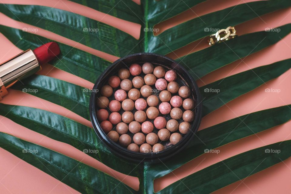 One round powder puff with pellets, red lipstick and a gold ring on a palm branch on a pink background, flat lay. Concept female cosmetics, beauty salon.