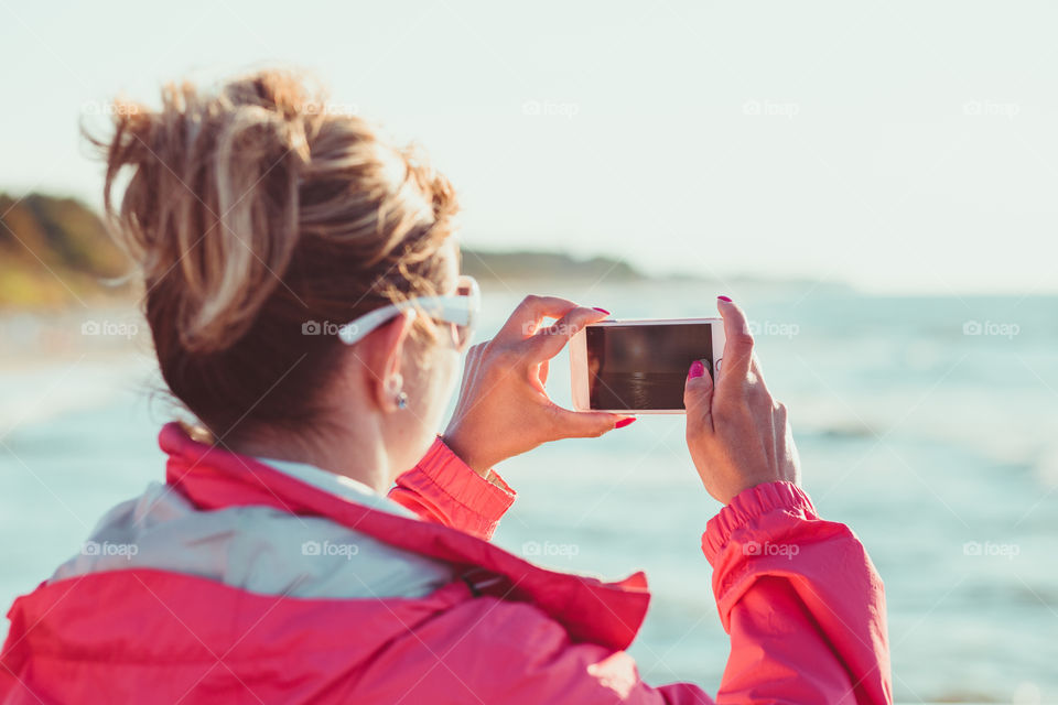 Young woman taking a photos using phone, looking at screen, standing outdoors, she is backlighted by sunlight with plain sky and sea in the background