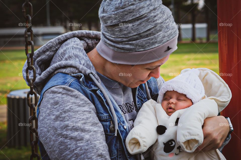 Newborn baby with mother in park on swing, newborn baby girl in big puffy panda suit, newborn baby’s first swing ride with mama, mother looking lovingly yay newborn child, newborn baby girl and mama 