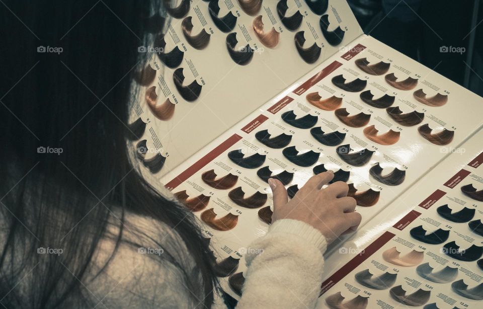 A young caucasian girl from the back sits on a chair in a hair salon holding a book with a hair palette for dyeing and chooses her favorite color with her finger, close-up side view. Concept of a hair salon, beauty salon, hairdresser services.