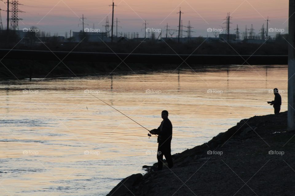 silhouette of a fisherman with a fishing rod near the river against the backdrop of city buildings. sunset