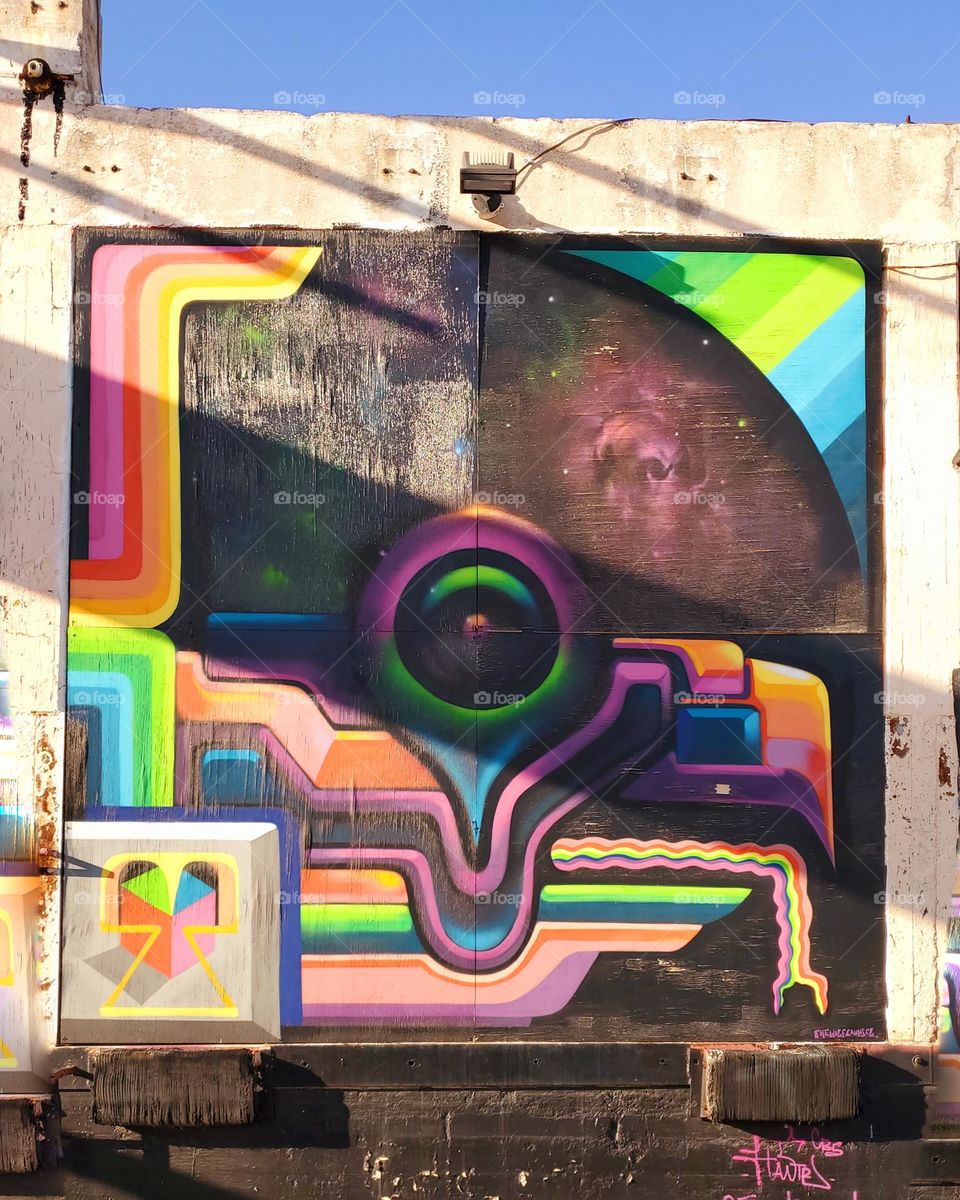 A bright and colorful mural with various hues of pink and diagnol shadows running across. It is located at an abandoned outdoor mural art gallery in the city.