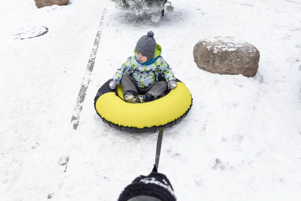 A small, carefree boy walks in the white snow in winter and rides a tubing in the park, near trees in the snow.