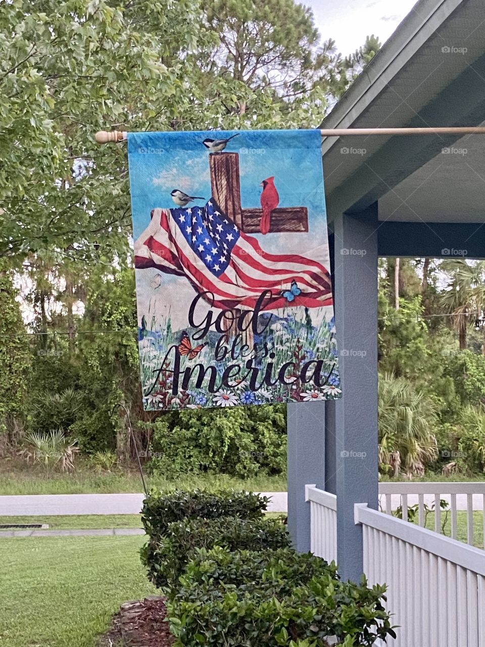 A decorative patriotic flag hanging on a flag pole from the front porch of a blue house with a white fence 