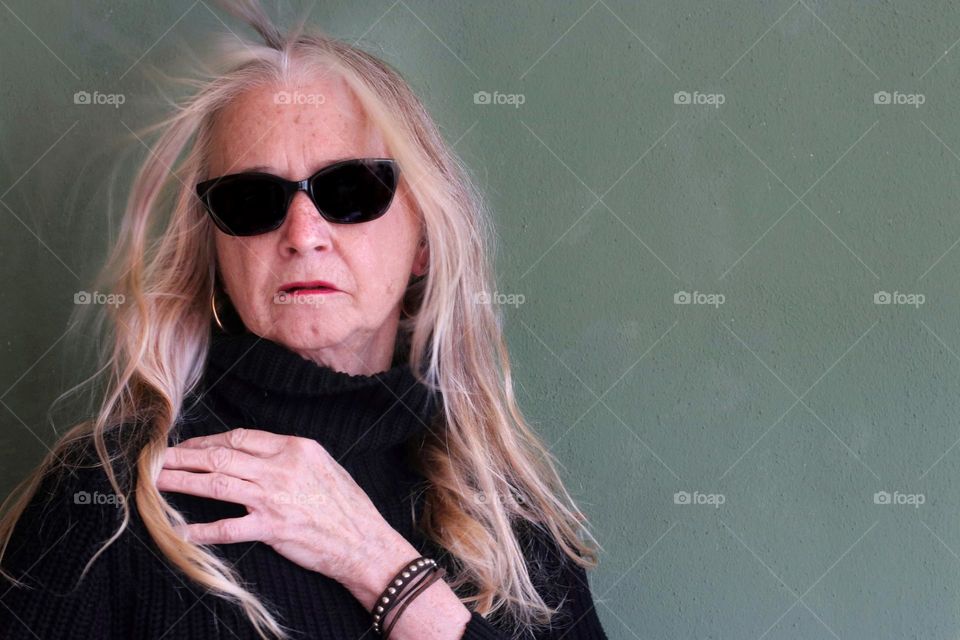 Portrait of an older woman with long blown blond hair, sunglasses and gold earrings against green background 