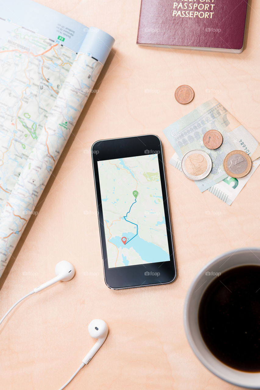 Smartphone with navigation map on screen, euro banknotes, paper map, passport, earphones on the wooden desk. Tourist essentials. Things related to travel