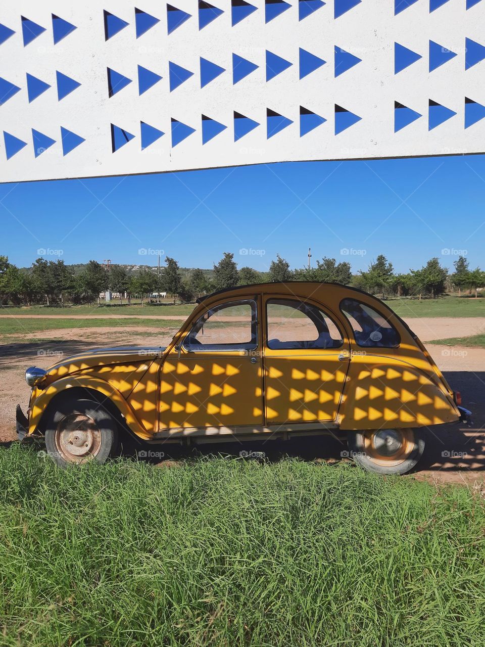 A Citroën 2CV in a field under a sign with triangular holes producing light reflections on the vehicle and creating a curious natural pattern.