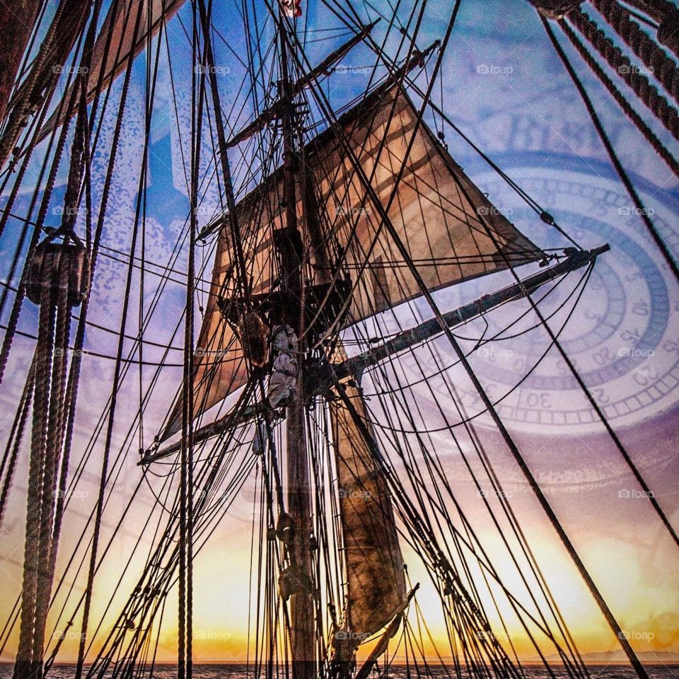 A mast and sail tower overhead this tall ship as it sails into the sun setting beneath the orange horizon. A nautical compass can be seen in the Sky’s above blended into the background of pirate ship in this sailors dreamscape.