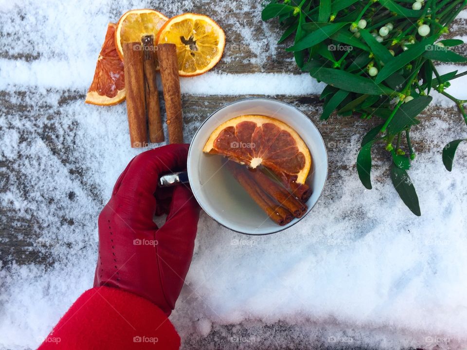 Woman’s hand wearing red glove holding cup of orange tea with mistletoe placed beside