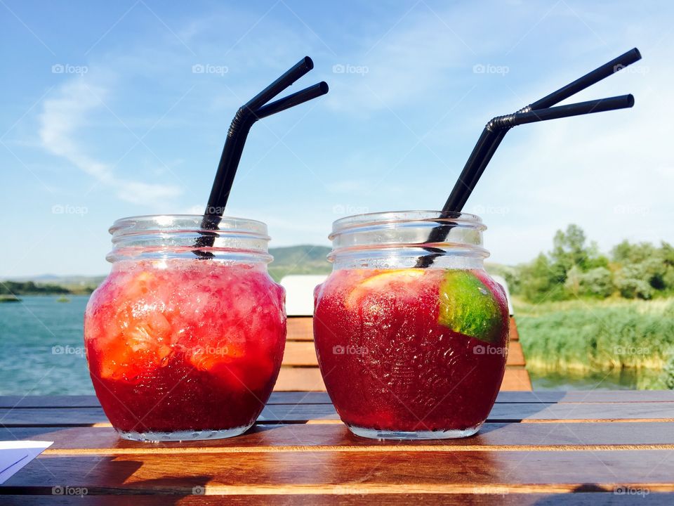 Two colourful glasses with strawberry lemonade on wooden table with lake in the background 