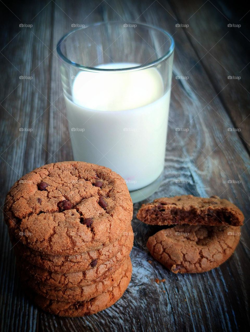 Chocolate chip cookies stacked on top of each other.  Nearby is a broken chocolate chip cookie.  In the background is a glass of milk.  Wooden background