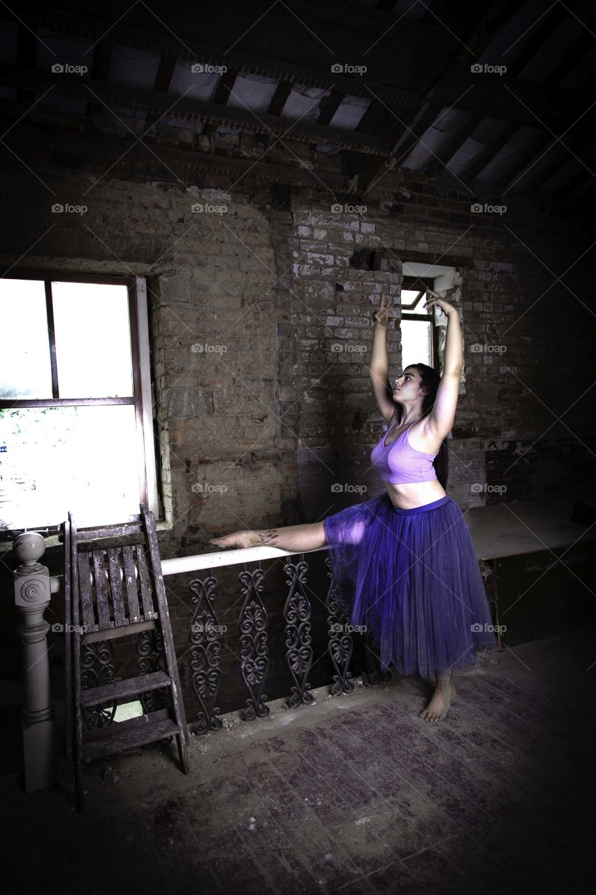 Girl dancing in a derelict building. Leg stretched on top of stair rail at the top of the stairs. Holding a dance pose. Step ladders leaning against hand rail. 
