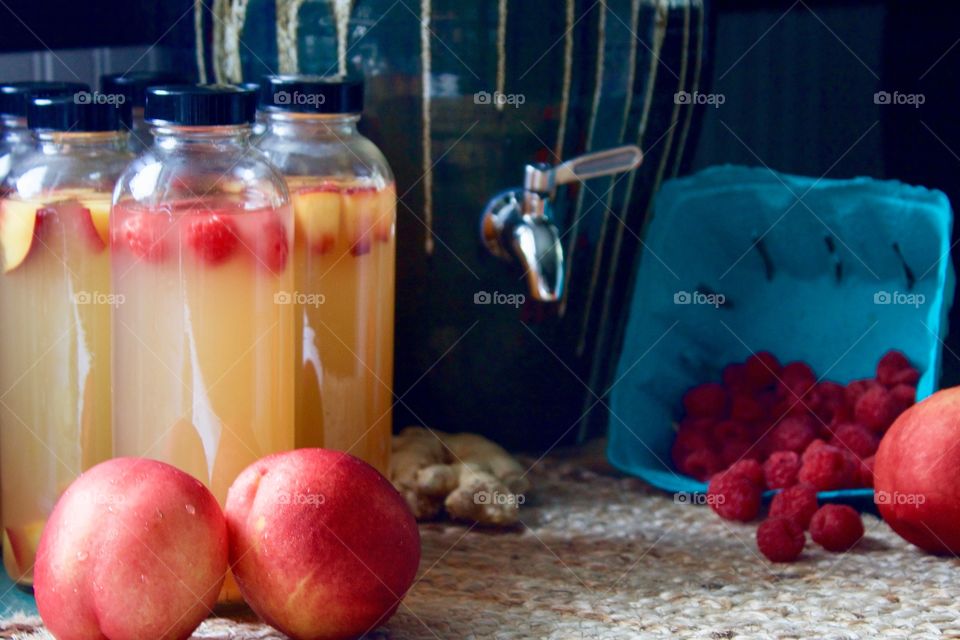 Kombucha, bottled for a second-ferment, flavored with nectarines, raspberries and ginger root slices on a natural fiber mat, whole nectarines and raspberries in a paper carton, stoneware kombucha crock with a stainless steel spigot in the background