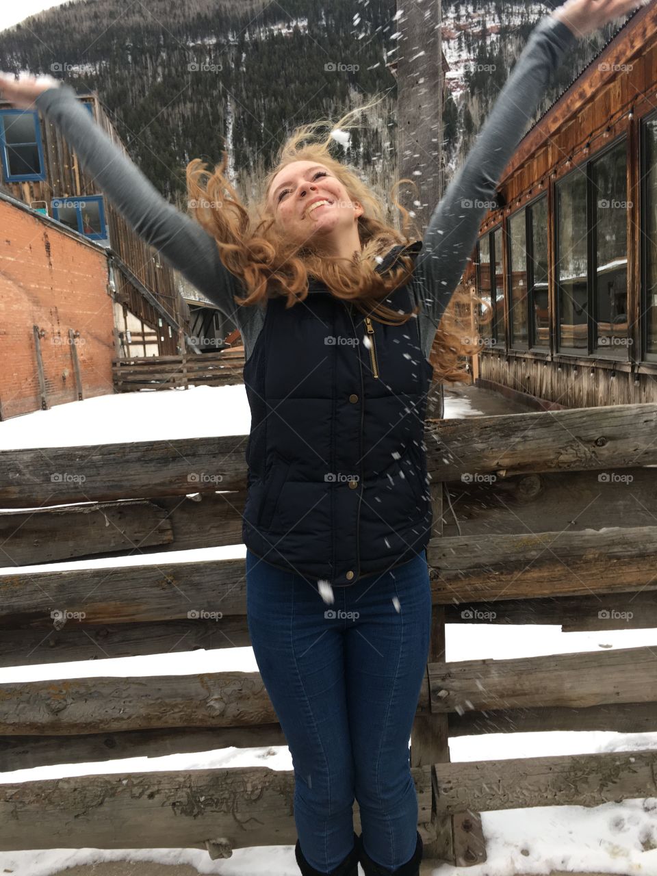 A young woman happily tosses snow in the air, as an appreciation of the beauty of overlooked commonalities. The dark wood behind her creates a dark contrast to the stark white snow. 