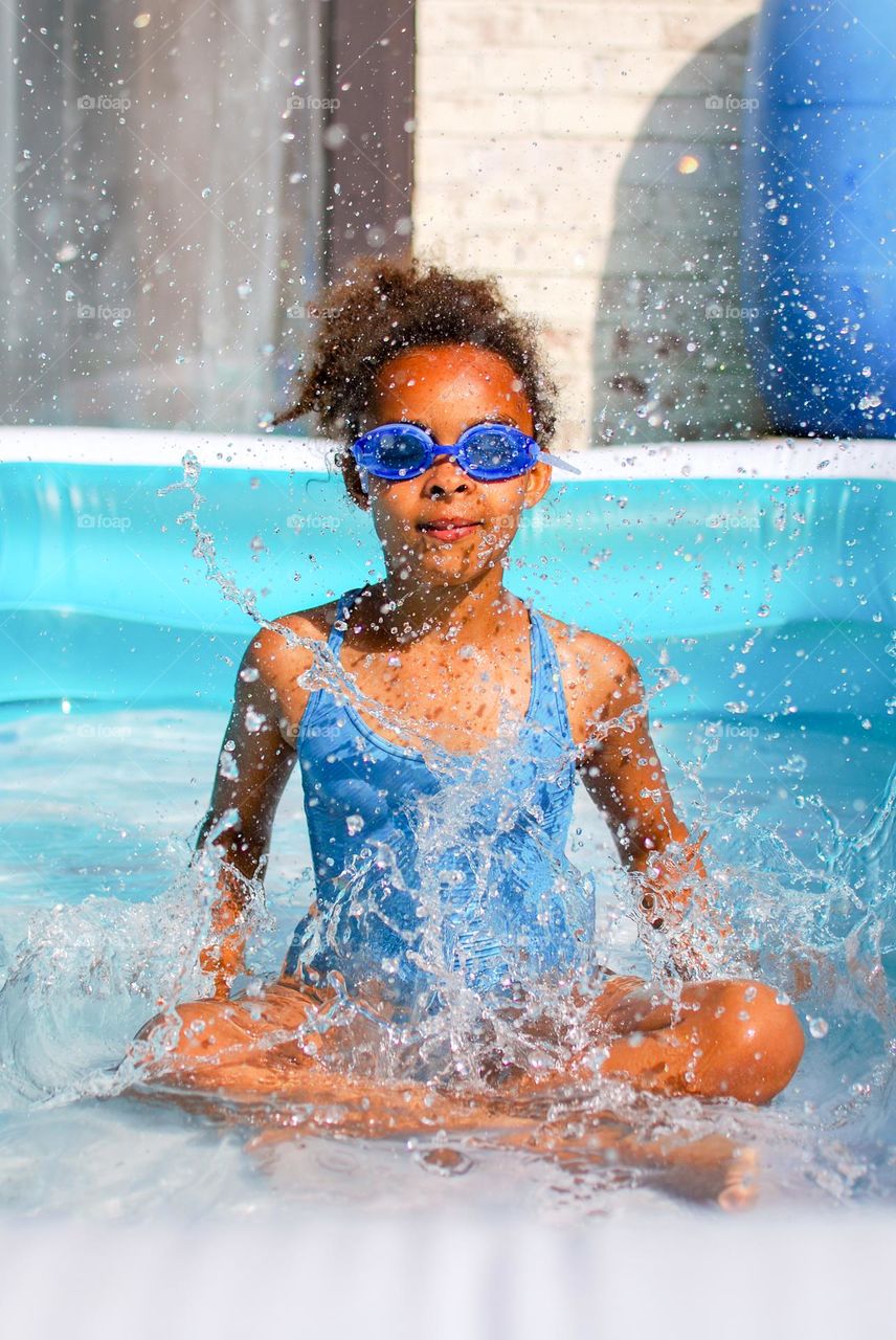 Girl of mixed race enjoying the refreshment of water in a swimming pool on a hot summer day, together with her little sister (family, fun, summer, water, blue, swimming suit, splash, hot, enjoy, play, outdoors)