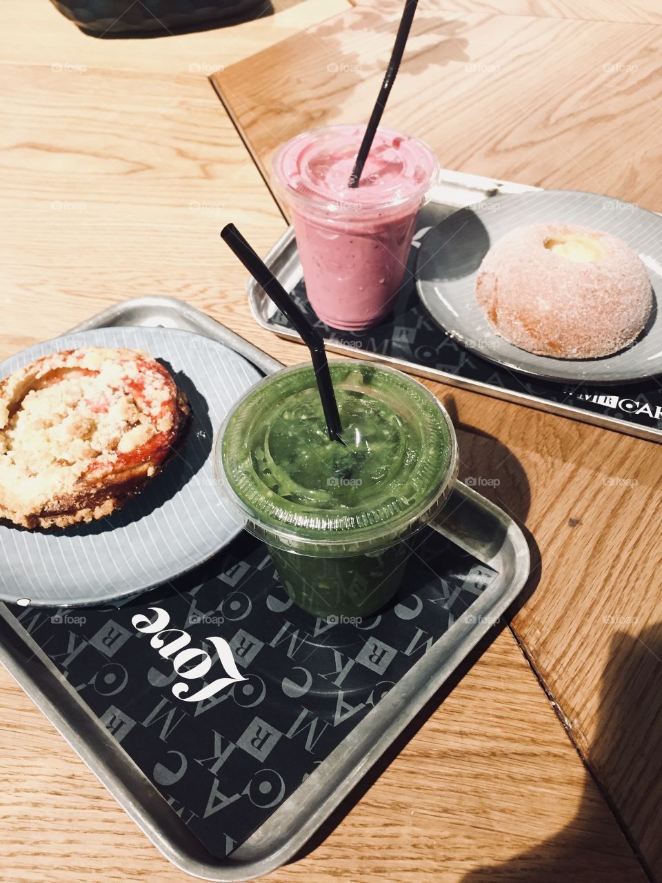 Colorful smoothies sit on trays next to freshly made pastries. A wooden table is beneath the two trays. 