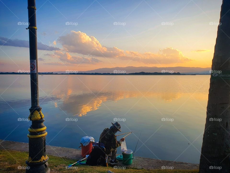 A man fishing at sunset on Phayao Lake in Phayao, Thailand. The reflection of the clouds and the colors made it a wonderful moment.