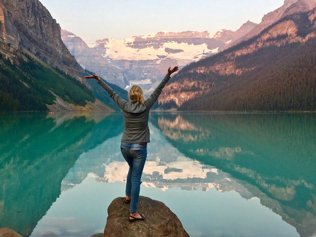 Woman standing on large rock in front of beautiful Lake Louise in Canada's Rocky Mountains with arms outstretched in praise and wonder 