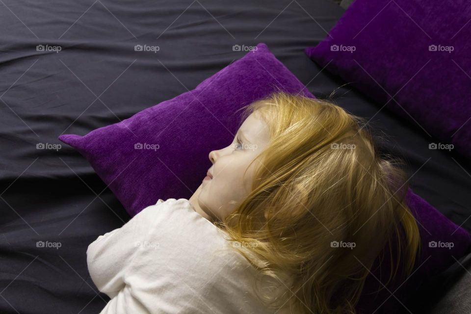 A small, red-haired girl is laughing at home on a gray sofa with purple pillows and a yellow background