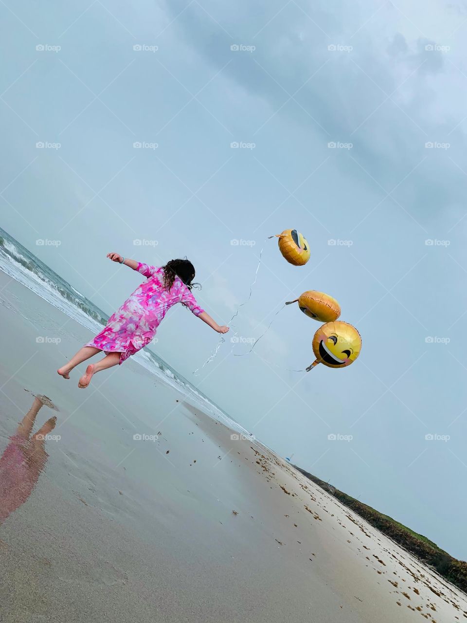 Little Girl Seems To Be Flying And Carried Up In The Air By The Three Helium Emoji’s Balloons On The Beach By The Ocean.