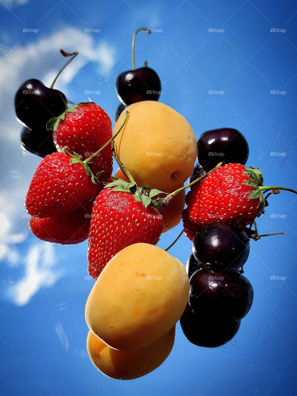 Summer fruit symphony. Fruits lie on a mirror surface: red strawberries, black cherries and yellow apricots. Fruits and a blue sky with white clouds are reflected from the mirror surface.