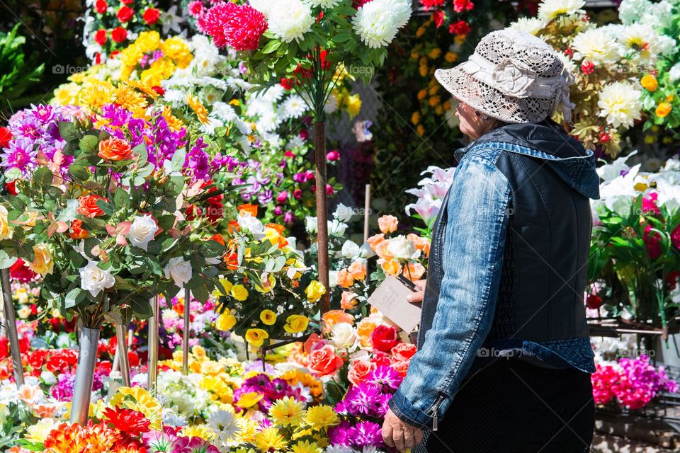 Old woman in blue coat and white hat standing in front of flower market