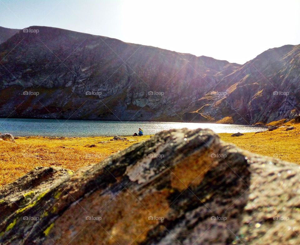 A beautiful view of Rila mountain in Bulgaria with a lake and hills and a clear sky over the green grass, a shining sun and rocks