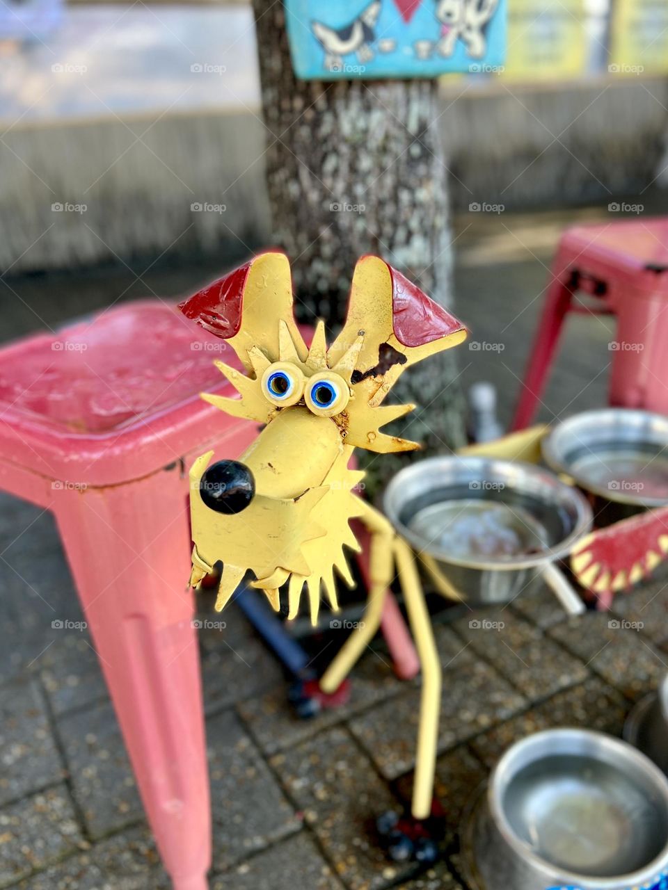 Yellow metal sculpture of a happy dog holds metal water bowls for passing dogs and birds to enjoy. On a city sidewalk by a cafe with red metal stools, next to a shade tree.