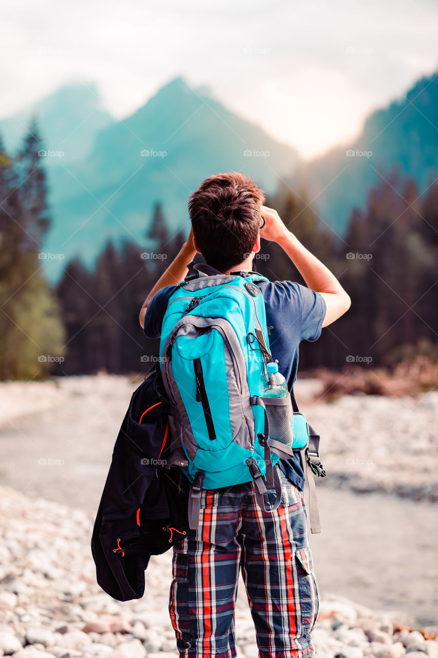 Young wanderer with backpack looks through a binoculars on mountains peaks, stands over a river. Boy spends a vacation in mountains, wandering with backpack, he is wearing sports summer clothes