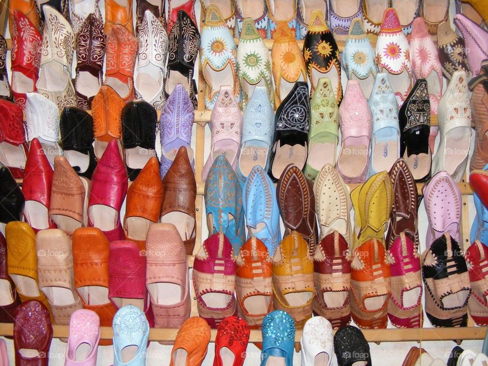 "A vibrant collection of traditional Moroccan slippers, known as 'babouche,' displayed on a wall. The shoes feature intricate handmade designs and a variety of bright colors, including red, orange, yellow, pink, blue, green, black, and white. The ima