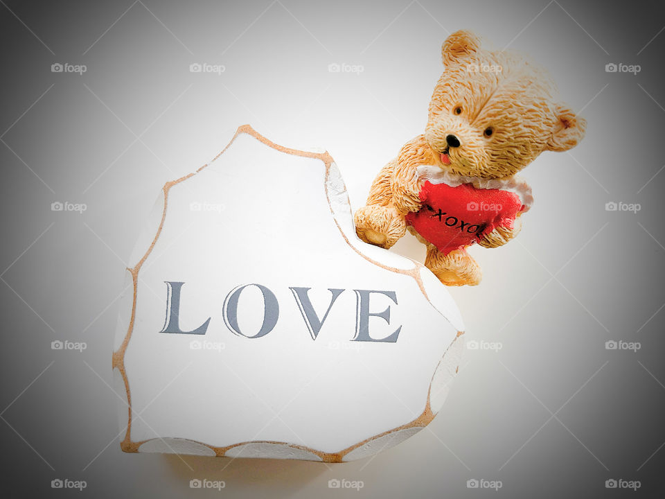 white wooden heart with the word LOVE wrote on it, mini teddy bear with hugs & kisses cushion in hands. White background.