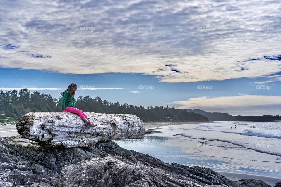 Girl sits on huge log that has washed onto rock outcropping of beach - a strange sight in Tofino, British Columbia, Canada 