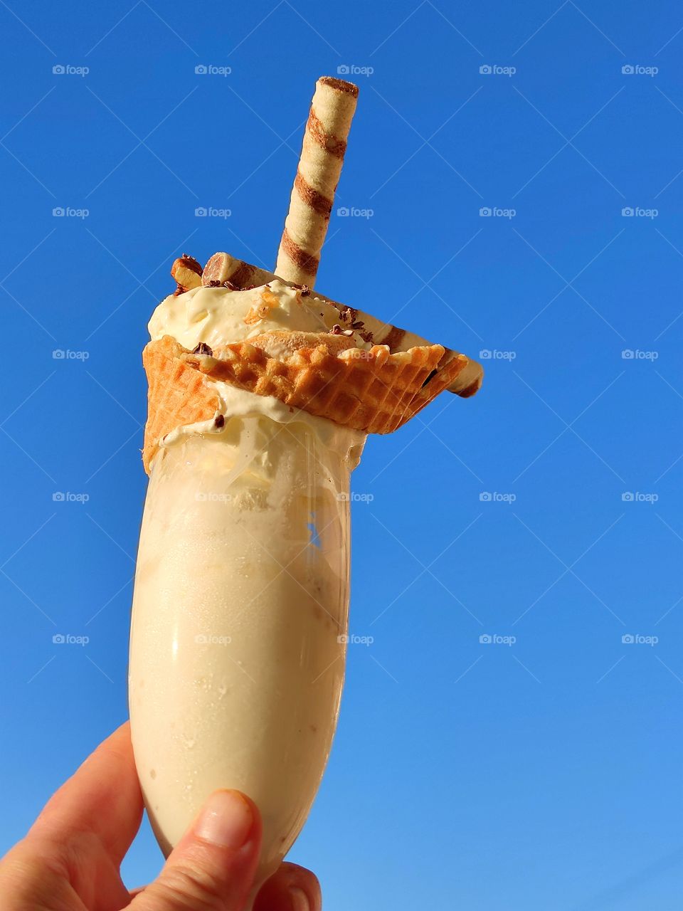 A woman's hand holds a glass of ice cream, the edge of which is wrapped with a waffle ribbon.  Ice cream sprinkled with chocolate chips and wafer rolls.  Sunny day.  Blue sky