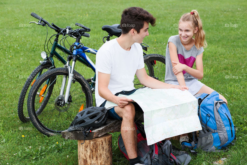 Young smiling boy and girl having a break sitting on bench during bike trip and planning a further part of trip with map equipped with helmets backpacks and bicycles