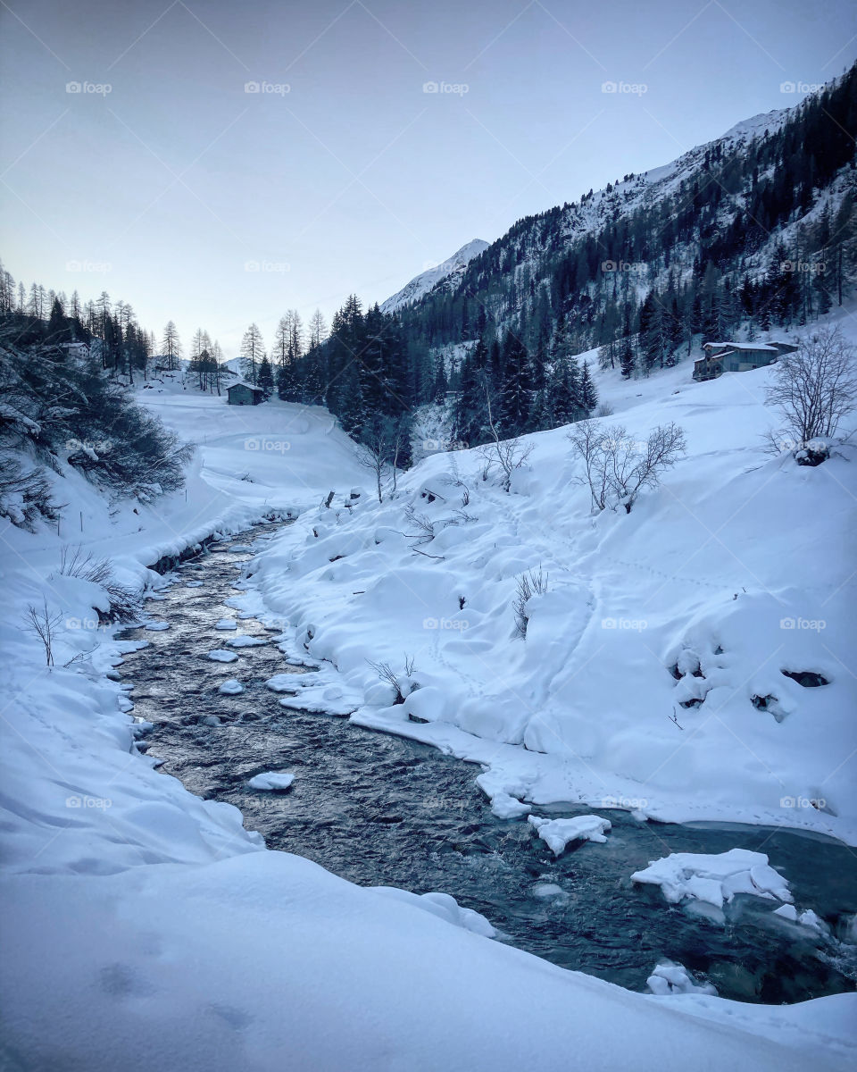 Winter landscape with river, snow covered mountain, remote place in the Swiss Alps, sunrise in a snowy Swiss valley, ice cold river reflecting the morning sky, cloudless winter day, Switzerland in winter