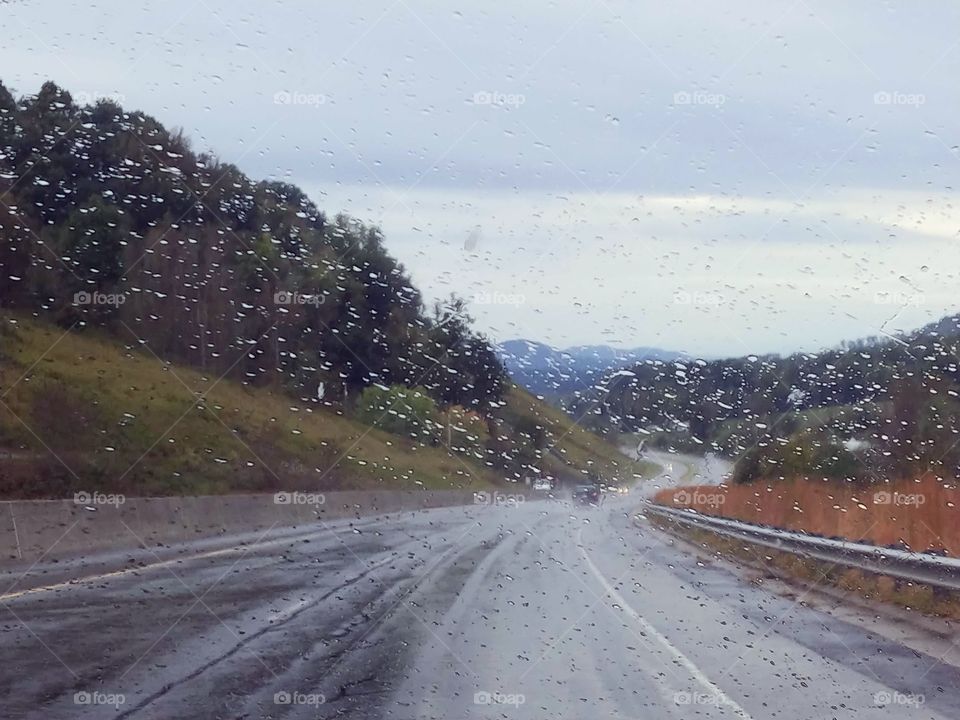 Raindrops on a windshield while driving through the mountains on an empty road with bright orange wild flowers on the side.