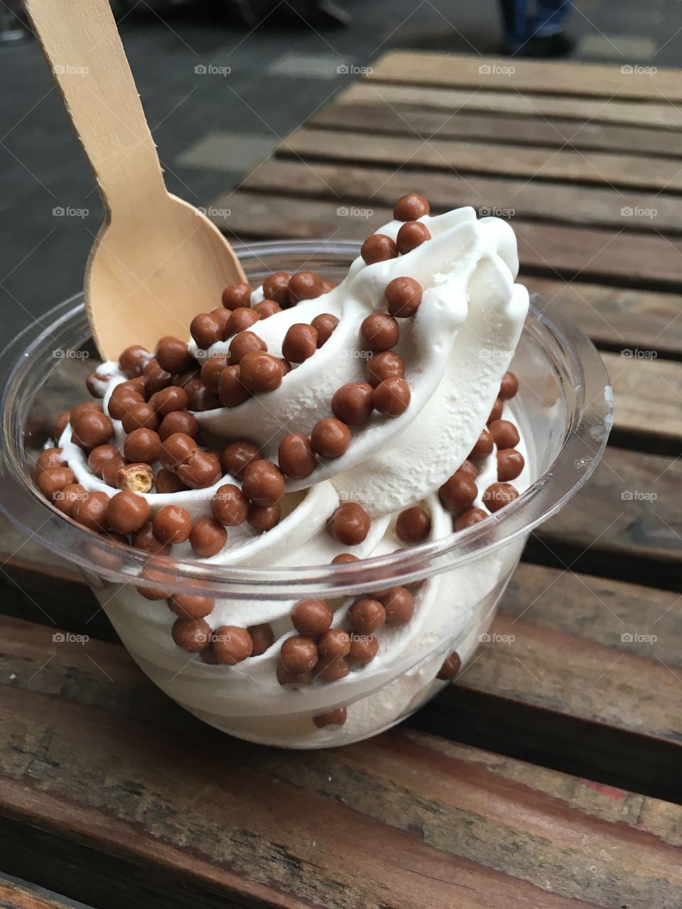 A vanilla soft serve ice cream in a plastic cup with little chocolate candies as a topping. On a wooden table with a wooden spoon 