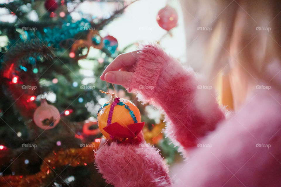 Young girl decorating Christmas tree, holding Christmas ball. Teenage blonde girl wearing pink sweater. Young girl celebrating Christmas at home
