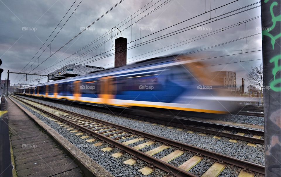 yellow and blue train at full speed 8n an urban environment on a railroad with high voltage cables and clouds above