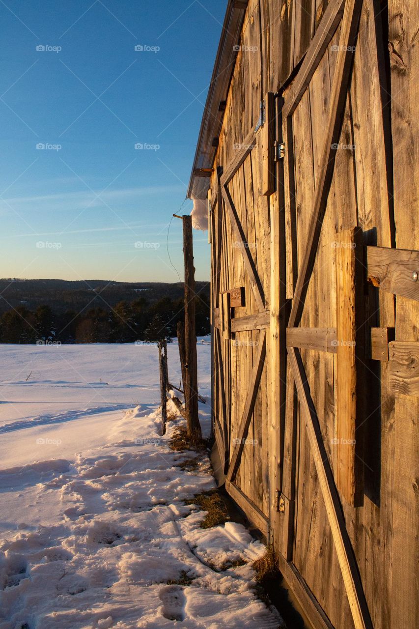 Bright warm wooden shed in the setting sunlight with snow covered ground, a mountainous background, and blue skies.