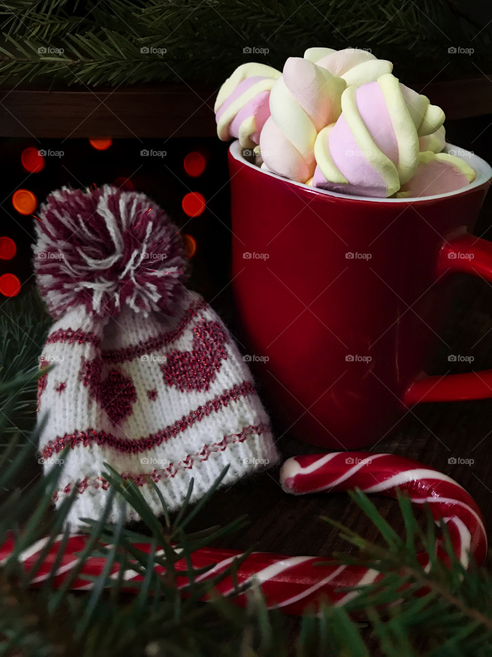 Red coffee cup with marshmallows, winter hat, red candy cane and Christmas tree branch in front of red night lights