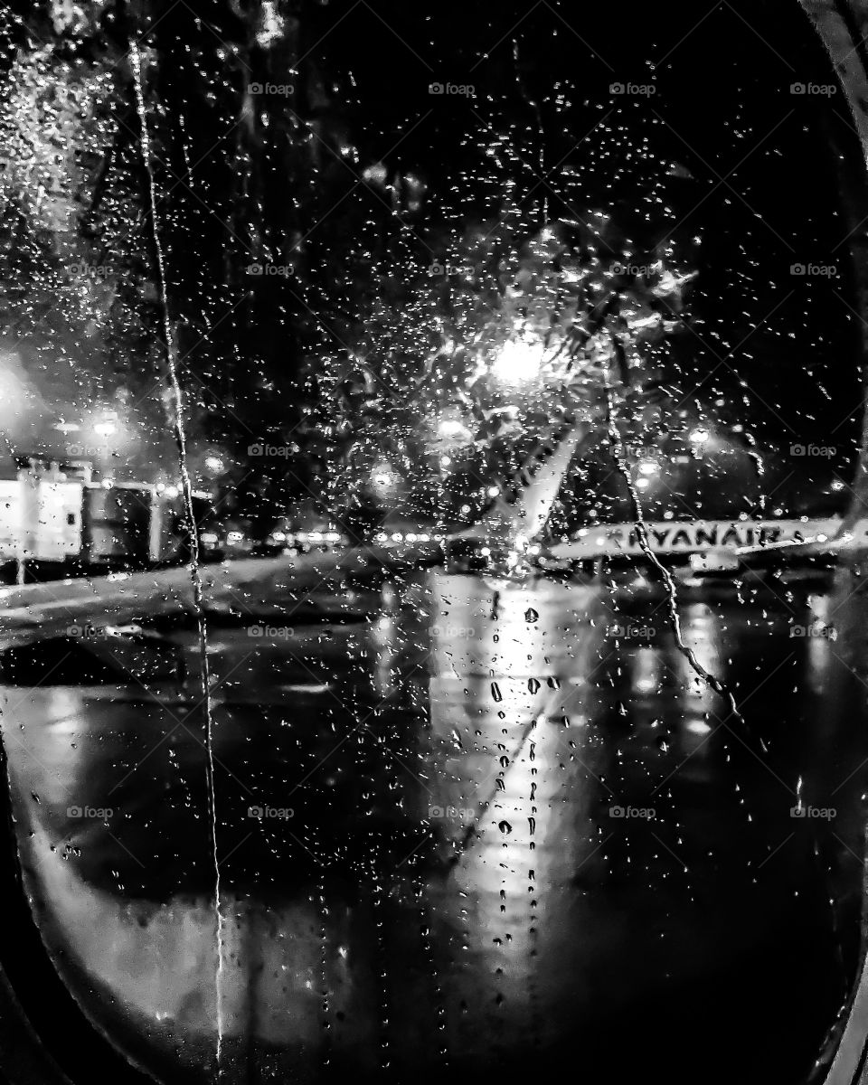 Black and white photograph of the view out of an aircraft window, with raindrops on the window on a dark gloomy night. 