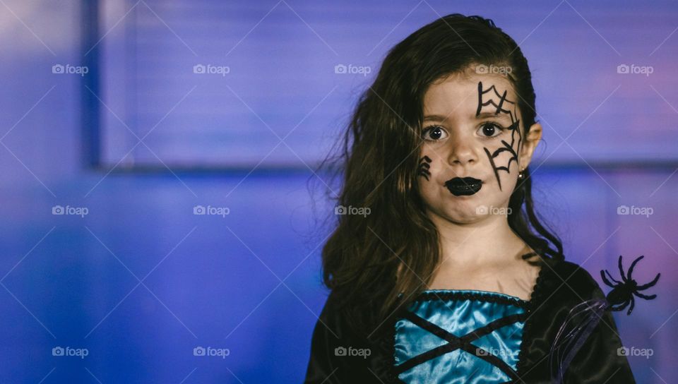 Portrait of a beautiful caucasian girl with big eyes, halloween makeup on her face, shoulder-length brown hair and in a witch dress stands on the right against the background of a neon wall, looking at the camera with copy space on the left, close-up