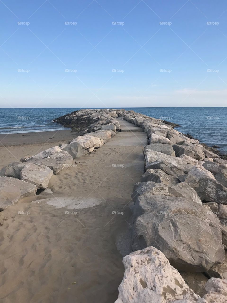 A road made of cement, a bit sandy above, filled with rocks on both sides , built on the beach . The sea is calm without many waves , the sky is light blue, with few clouds in the distance