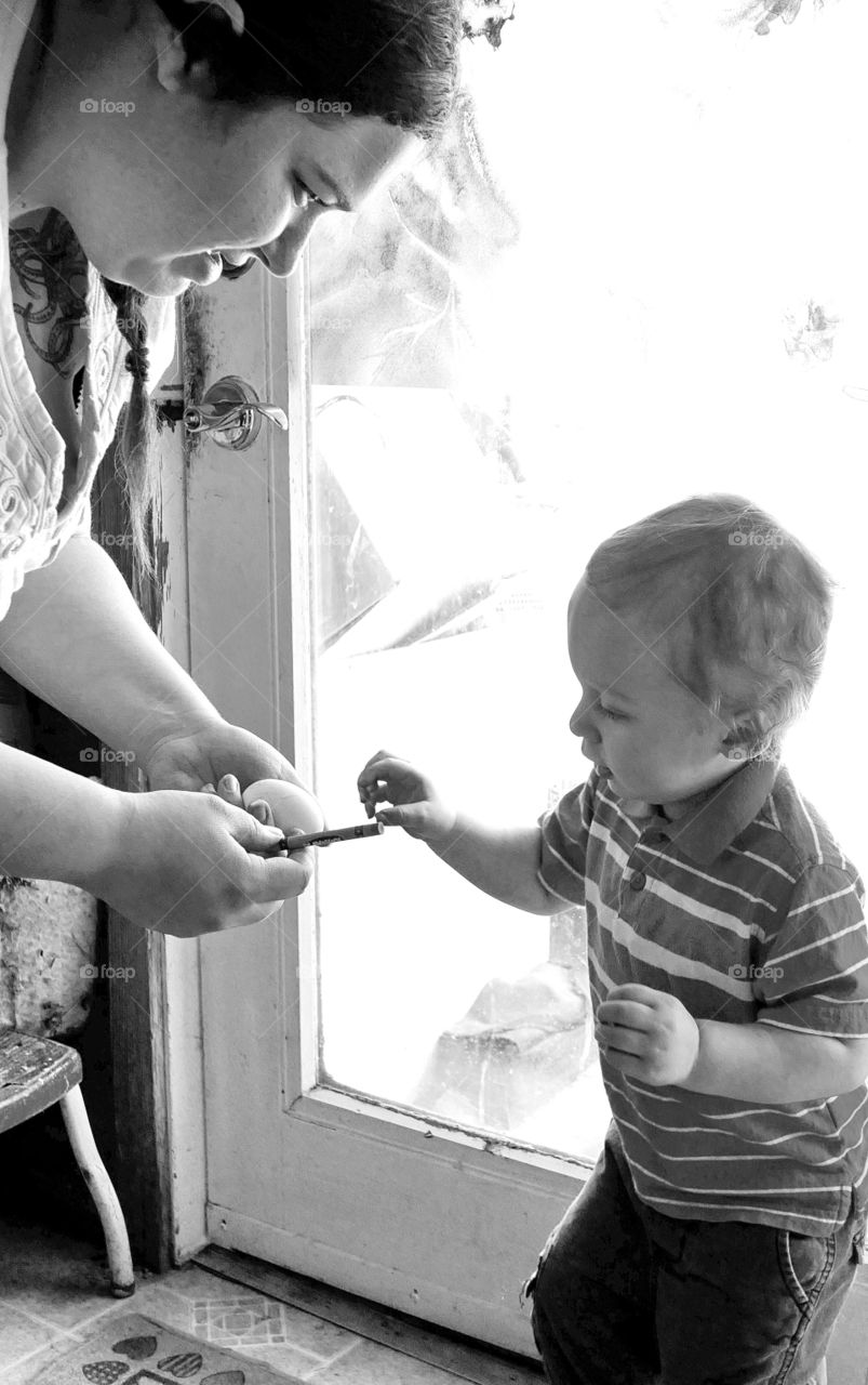 A mother hands her toddler son a crayon and an egg to draw on for his second Easter. 