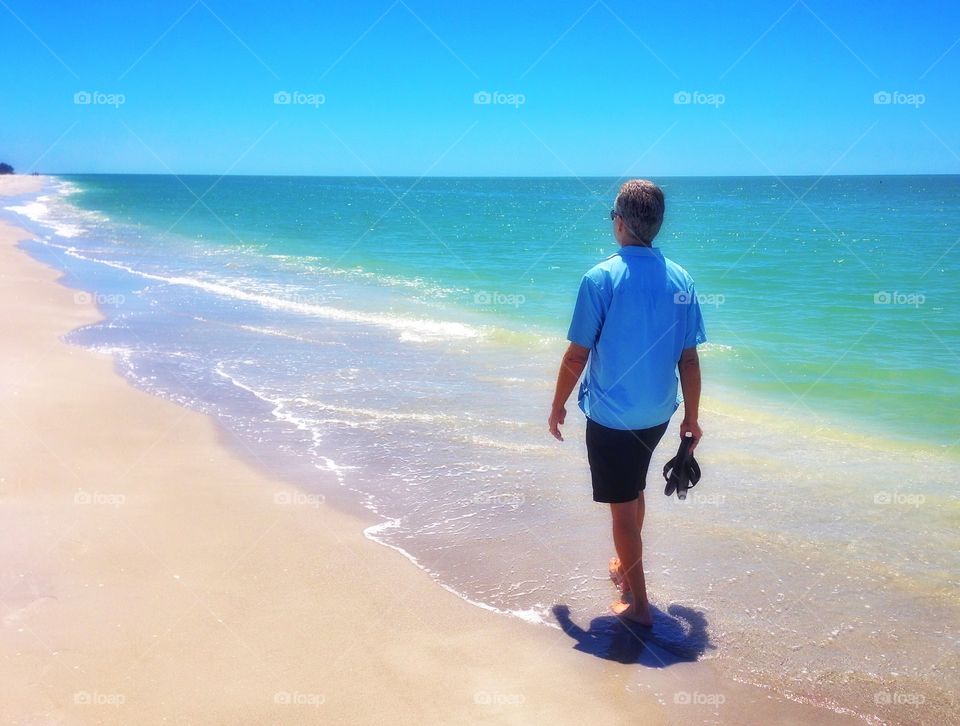 A man strolls up the sandy beach toward new horizons as the warm ocean waves lap at his feet