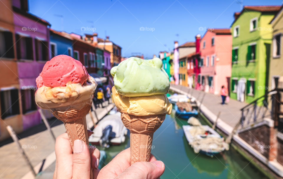 Two hands holding colorful ice cream in Burano island in region of Venice, Italy. Colorful houses and boats.