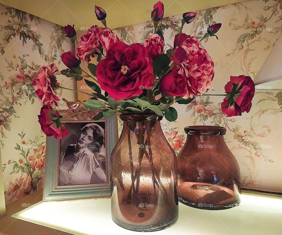 There are dark glass bottles on the shelf.  In one bottle there are roses in the form of blossoming flowers and buds.  Roses in all shades of red.  Green leaves break through between the roses.  In the background is a framed black and white photograp